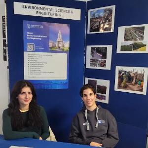 2 students sitting at desk smiling with images on a backdrop and course information booklets on table